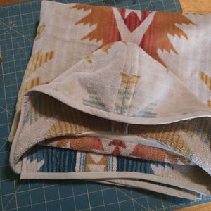 Puede incluir: Una toalla con capucha beige doblada con un estampado de inspiraci&oacute;n suroeste en tonos naranja, rojo y azul. La toalla est&aacute; hecha de material suave y tiene un ribete blanco. Se ve una taza amarilla en la esquina.