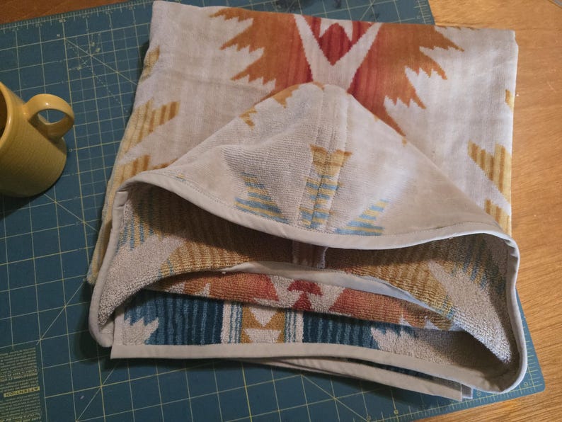 Puede incluir: Una toalla con capucha beige doblada con un estampado de inspiraci&oacute;n suroeste en tonos naranja, rojo y azul. La toalla est&aacute; hecha de material suave y tiene un ribete blanco. Se ve una taza amarilla en la esquina.