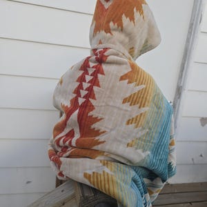 Puede incluir: Una manta con capucha con un dise&ntilde;o de inspiraci&oacute;n suroeste. La manta presenta una base color crema con patrones geom&eacute;tricos en naranja, rojo, amarillo y azul. La capucha est&aacute; levantada y la manta est&aacute; sobre una barandilla de madera.