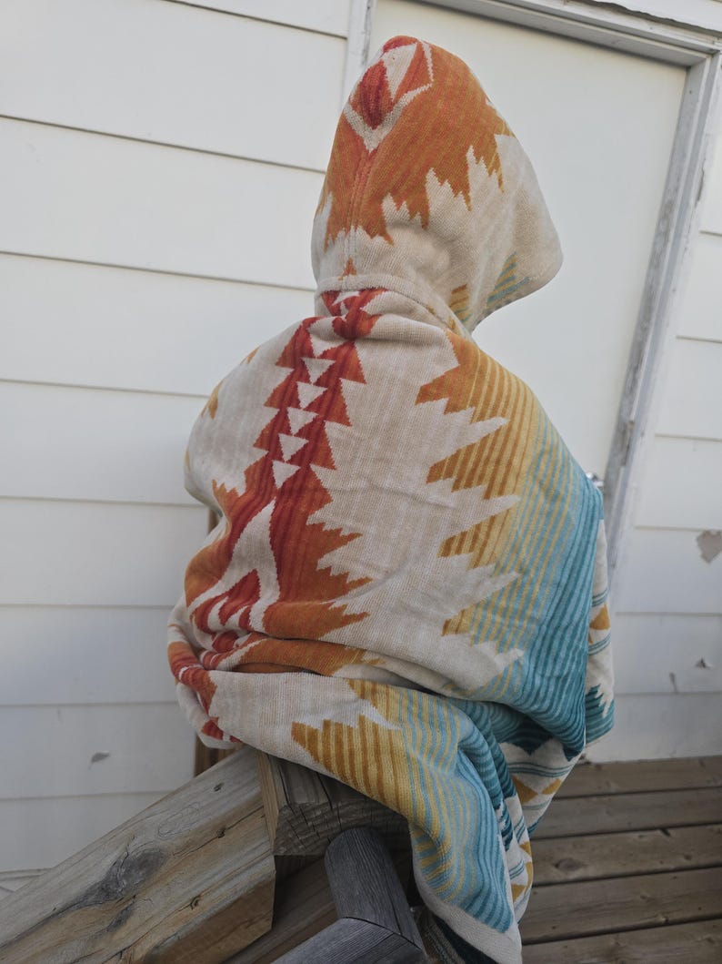 Puede incluir: Una manta con capucha con un dise&ntilde;o de inspiraci&oacute;n suroeste. La manta presenta una base color crema con patrones geom&eacute;tricos en naranja, rojo, amarillo y azul. La capucha est&aacute; levantada y la manta est&aacute; sobre una barandilla de madera.