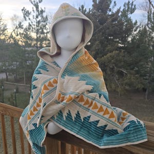 Puede incluir: Una toalla con capucha con un patr&oacute;n geom&eacute;trico en colores turquesa, amarillo y crema. La toalla est&aacute; colocada sobre una cabeza de maniqu&iacute; en una barandilla de madera, con un fondo natural al aire libre.