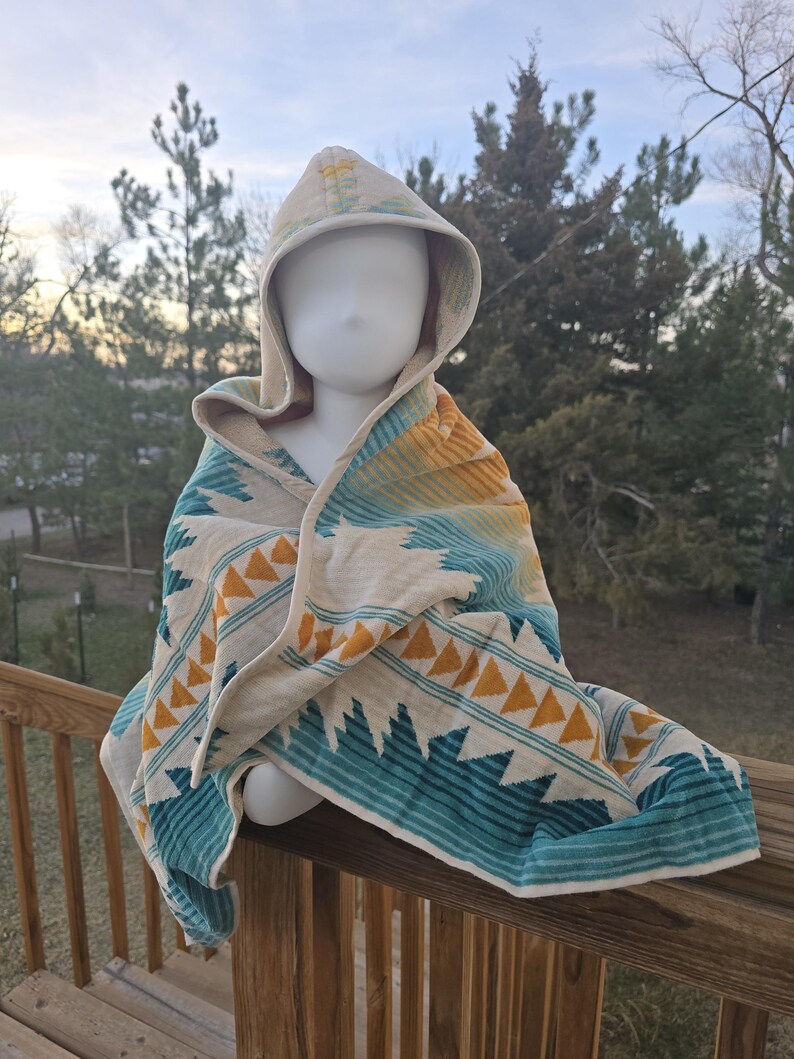 Puede incluir: Una toalla con capucha con un patr&oacute;n geom&eacute;trico en colores turquesa, amarillo y crema. La toalla est&aacute; colocada sobre una cabeza de maniqu&iacute; en una barandilla de madera, con un fondo natural al aire libre.
