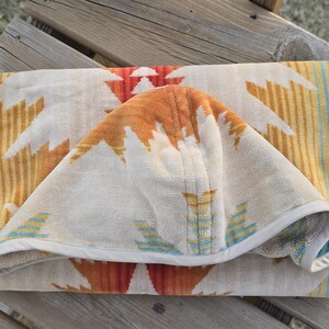 Puede incluir: Toalla de playa doblada con un estampado del suroeste. La toalla presenta un fondo crema con dise&ntilde;os geom&eacute;tricos en naranja, amarillo y blanco. Los bordes est&aacute;n ribeteados con un borde blanco y rayas azules.