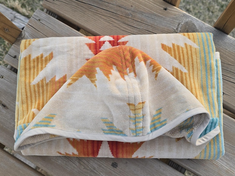 Puede incluir: Toalla de playa doblada con un estampado del suroeste. La toalla presenta un fondo crema con dise&ntilde;os geom&eacute;tricos en naranja, amarillo y blanco. Los bordes est&aacute;n ribeteados con un borde blanco y rayas azules.