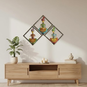 May include: Three colorful metal wall art pieces depicting figures in traditional attire, each framed in a black diamond shape. The figures are adorned with turbans and vibrant clothing, with a wooden cabinet and plant in the foreground.