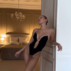K&ouml;nnte beinhalten: Ein schwarzer Body mit tiefem V-Ausschnitt und transparenten Tr&auml;gern. Das einteilige Kleidungsst&uuml;ck hat ein Schl&uuml;ssellochdetail an der Brust. Der Hintergrund zeigt ein Schlafzimmer mit Bett und Kronleuchter.