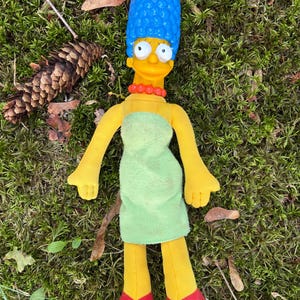 May include: A plush doll of Marge Simpson, featuring her signature blue beehive hairstyle, yellow skin, and a green dress. The doll has red shoes and a red bead necklace. The doll is lying on a bed of green moss.