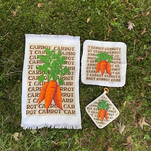 May include: A set of four white kitchen textiles with a carrot theme. The set includes a fringed towel, a square hot pad, and a diamond-shaped pot holder, all featuring orange carrots and green tops. The word "CARROT" is repeated on each item.