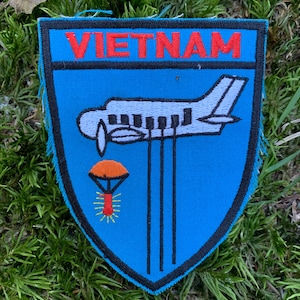 Puede incluir: Un parche bordado azul y negro con la palabra "VIETNAM" en letras rojas. Un avión blanco con un paracaídas y una bengala roja se representa en el parche.