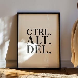 May include: A framed print featuring the text "CTRL. ALT. DEL." in a bold, black font. The print has a cream-coloured background and a black frame. The artwork is leaning against a white wall, with a wooden chair and a beige blanket in the background.