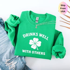May include: A bright green sweatshirt with the text "DRINKS WELL WITH OTHERS" and a white four-leaf clover graphic. A silver necklace with a small pendant is on the sweatshirt. The sweatshirt is folded next to a pair of blue jeans.