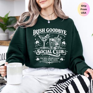 May include: A dark green sweatshirt with the text "IRISH GOODBYE SOCIAL CLUB" in white. The design includes illustrations of cocktails and shamrocks. The sweatshirt is a casual garment, suitable for everyday wear.