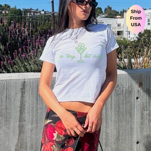 May include: White t-shirt with a green cocktail glass and text that reads "a tiny bit irish". The shirt is short-sleeved and has a crew neck. The design is printed in green. The model is wearing sunglasses and a floral skirt.