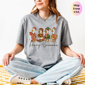 May include: A gray t-shirt featuring a whimsical illustration of four geese dressed in festive Christmas attire. The geese are depicted with Santa hats, scarves, and holding holiday items. The text "Merry Goosmas" is written below the geese.