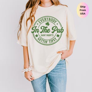 May include: Cream-colored t-shirt with a green circular graphic that reads "Everybody In The Pub, Saint Paddy's, Gettin' Tipsy" with shamrock accents. The shirt is worn with blue jeans.