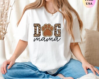 May include: White t-shirt with the words "DOG mama" in a black script font. The word "DOG" is in a leopard print font with a brown paw print in the center. The shirt is made of a soft material.