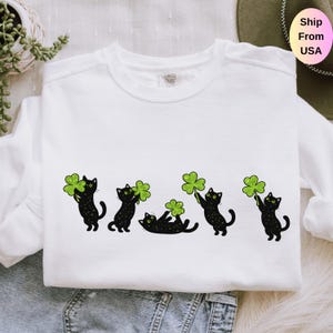 May include: White sweatshirt featuring a playful design of black cats holding green shamrocks. The cats are in various poses, adding a whimsical touch to the St. Patrick's Day themed apparel. The sweatshirt is made of a soft material.