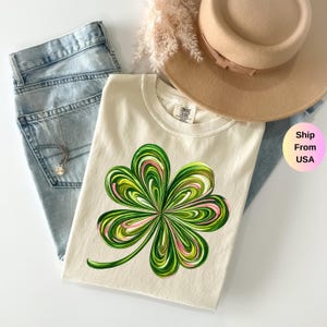 May include: Cream-colored t-shirt featuring a vibrant, abstract shamrock design in shades of green and pink. The shirt is paired with light-wash denim jeans and a tan hat. A delicate necklace is visible.