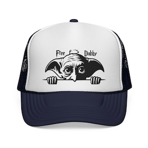 May include: A navy blue and white trucker hat featuring a black and white graphic of a creature with the text "Free Dobby". The hat has a mesh back and a curved brim.