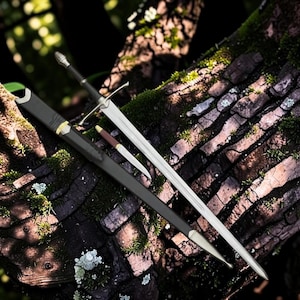 May include: A long steel sword with a black grip and a silver pommel, accompanied by a matching black scabbard with silver accents. A smaller dagger with a brown handle and a silver blade is positioned between the sword and the scabbard.