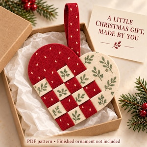 May include: A woven heart-shaped ornament made of red felt with white dots and cream felt with green leaf patterns. The ornament has a matching red felt hanging loop and is presented in a brown box with white tissue paper and a card reading A LITTLE CHRISTMAS GIFT, MADE BY YOU.