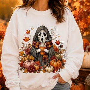 May include: A white long-sleeve sweatshirt featuring a graphic of a hooded figure in a dark cloak and plaid scarf holding a pumpkin-themed coffee cup, surrounded by colorful autumn leaves, pinecones, and various pumpkins in shades of orange.