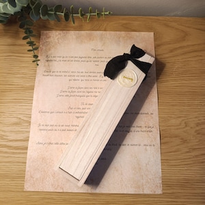 Puede incluir: Una caja de madera clara con una cinta negra y un sello de cera, sobre papel de estilo vintage con texto en francés. La caja es rectangular y tiene un diseño sencillo. El sello de cera es dorado.