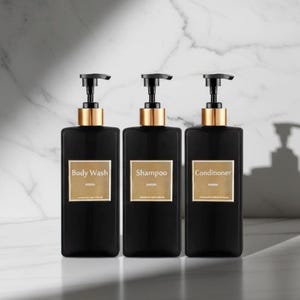 Puede incluir: Tres botellas negras cuadradas con detalles dorados y bombas negras. Cada botella tiene una etiqueta dorada con el texto: "Body Wash", "Champú" y "Acondicionador". Las botellas están dispuestas en fila sobre un fondo de mármol.