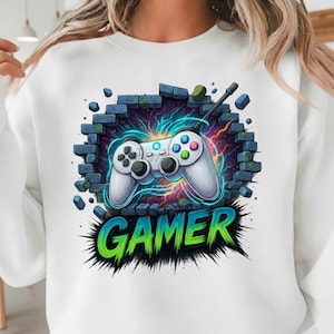 May include: White sweatshirt featuring a graphic of a video game controller breaking through a brick wall. The word "GAMER" is displayed in green and blue, with a lightning effect in the background.