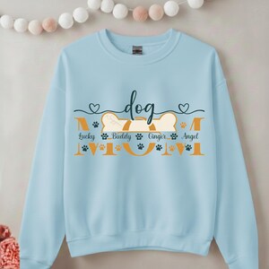 May include: Light blue sweatshirt with the word "dog" in cursive, a bone graphic, and the words "Mom" and the names of dogs. The design includes paw prints and hearts. The sweatshirt is a crew neck.