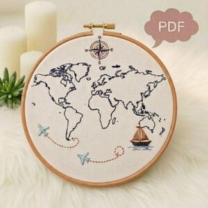 May include: Embroidered world map in a wooden hoop. The map is outlined in dark blue thread, with a compass rose at the top. Two airplanes and a sailboat are stitched on the map, with dotted lines indicating travel routes. The text "PDF" is in a speech bubble.