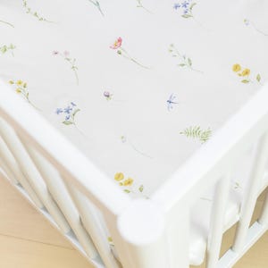 May include: White crib with a floral patterned sheet. The sheet features watercolor illustrations of wildflowers in shades of pink, yellow, blue, and green. The crib is made of white wood.