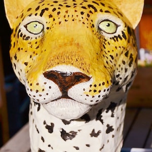 May include: A close-up of a papier-m&acirc;ch&eacute; sculpture of a leopard's head. The sculpture is painted in shades of yellow, brown, and black to resemble a leopard's fur. The sculpture has a realistic appearance, with detailed features such as the leopard's eyes, nose, and whiskers.