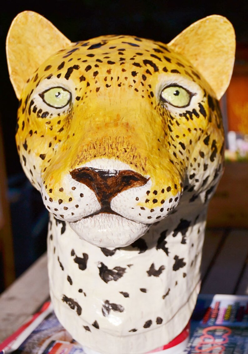 May include: A close-up of a papier-m&acirc;ch&eacute; sculpture of a leopard's head. The sculpture is painted in shades of yellow, brown, and black to resemble a leopard's fur. The sculpture has a realistic appearance, with detailed features such as the leopard's eyes, nose, and whiskers.
