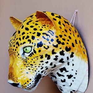 May include: A paper mache leopard head sculpture with black and yellow spots, painted white around the mouth and nose. The sculpture has green eyes and is hanging on a wall.