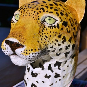 May include: A close-up of a papier-m&acirc;ch&eacute; leopard head sculpture. The sculpture is painted with realistic details, including black spots on a yellow and white background. The sculpture has a textured surface and is painted with green eyes.