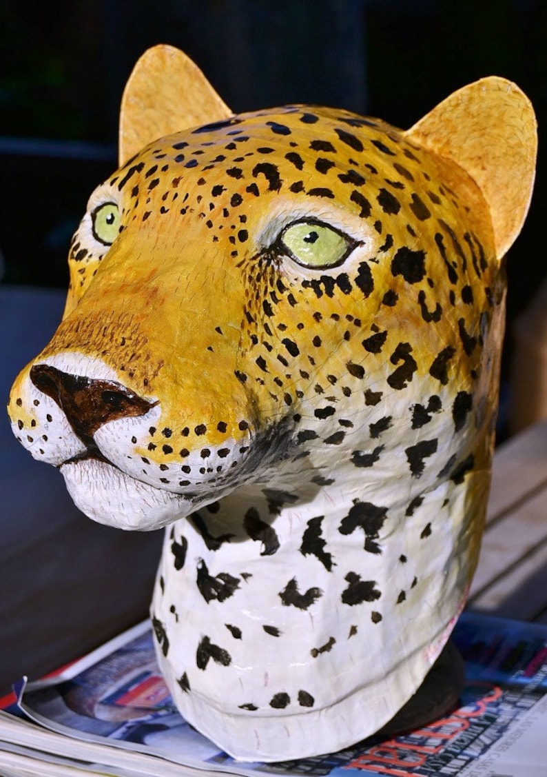 May include: A close-up of a papier-m&acirc;ch&eacute; leopard head sculpture. The sculpture is painted with realistic details, including black spots on a yellow and white background. The sculpture has a textured surface and is painted with green eyes.