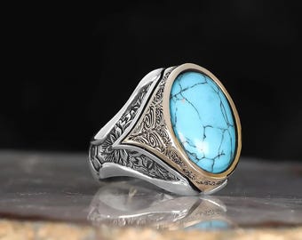 May include: A silver ring with an oval, light blue turquoise stone. The ring has a gold-colored bezel and ornate, engraved detailing on the band. The ring is displayed on a reflective surface.