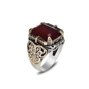 Op de afbeelding: Een zilveren ring met een grote, rechthoekige, dieprode edelsteen. De ring heeft sierlijke gouden en zwarte details rond de steen en op de band, met een gedetailleerd filigraanontwerp aan de zijkanten.