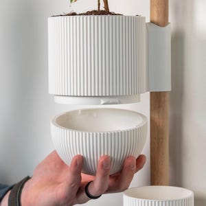 May include: A white, ribbed ceramic plant pot with a small plant. The pot is suspended by a white bracket attached to a wooden pole. A matching bowl is held in a hand below. The design is modern and minimalist.