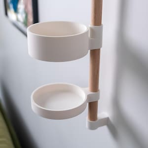 May include: A white, two-tiered wall-mounted organiser with a wooden pole. The organiser has a round, open-top container on top and a smaller, shallow tray below. The wooden pole is attached to the wall with white brackets.