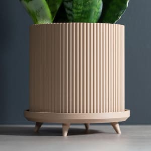 May include: A beige, cylindrical planter with vertical ribbed detailing. The planter sits on a matching saucer supported by three small, conical legs. Green plant leaves are visible inside the planter. A modern, decorative item for home decor.