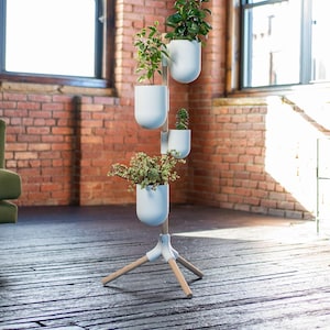 May include: A white, multi-tiered plant stand with four attached planters, each holding greenery. The stand has a tripod base with wooden legs and a white frame. The background features a brick wall and wooden floor.