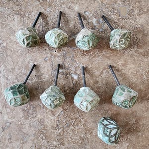 May include: A set of nine cabinet knobs with a faceted, geometric shape. Each knob features a weathered, sage green and white finish with a raised, grid-like pattern. Seven of the knobs have a threaded metal bolt extending from the back.