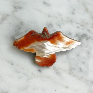 May include: A bird-shaped brooch carved from iridescent shell material, featuring a smooth, polished surface with a gradient of deep orange and reddish-brown tones transitioning into a pearlescent white centre across the wings and body.