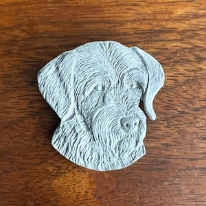 May include: A gray, sculpted dog head, likely a breed like a Wirehaired Pointing Griffon, with detailed fur texture. The dog's face is centered, with visible eyes, nose, and ears. The sculpture is set against a wooden background.