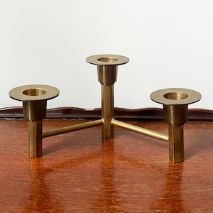 May include: A brass candle holder with three circular candle cups. The candle holder has a triangular base and is set on a wooden surface. The background is a plain white wall.