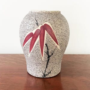 Może przedstawiać: Mały, zaokrąglony wazon ceramiczny o nakrapianej, jasnoszarej i beżowej teksturowanej powierzchni. Z przodu wazonu znajduje się ręcznie malowany wzór ciemnoczerwonych liści bambusa oraz cienkich, czarnych gałązek przypominających tusz.