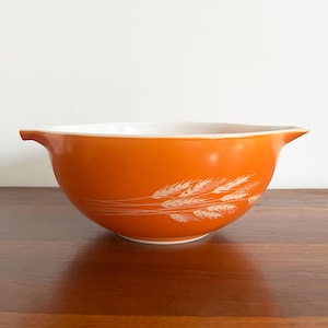 May include: Orange mixing bowl with a white interior and two small handles. The exterior features a white wheat pattern. The bowl is made of glass and sits on a wooden surface.