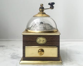 May include: An antique coffee grinder with a wooden base and brass accents. The grinder features a clear, domed top and a small drawer. The year "1865" is displayed on the front. The handle is black.